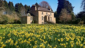 A large stone building with a symmetrical facade, a central entrance with steps, and a surrounding garden with yellow flowers. - 2 Bed in Beauly (oc-78962) (Windhill)