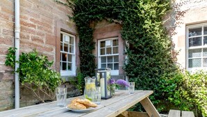 A wooden picnic table with a plate of croissants, a pitcher, and glasses, set against a brick wall with ivy and a window. - 2 Bed in Beauly (oc-78962) (Windhill)