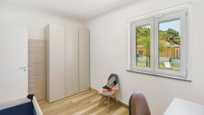 A room with a wooden floor, a white wardrobe, a colorful chair, and a window with a view of greenery.