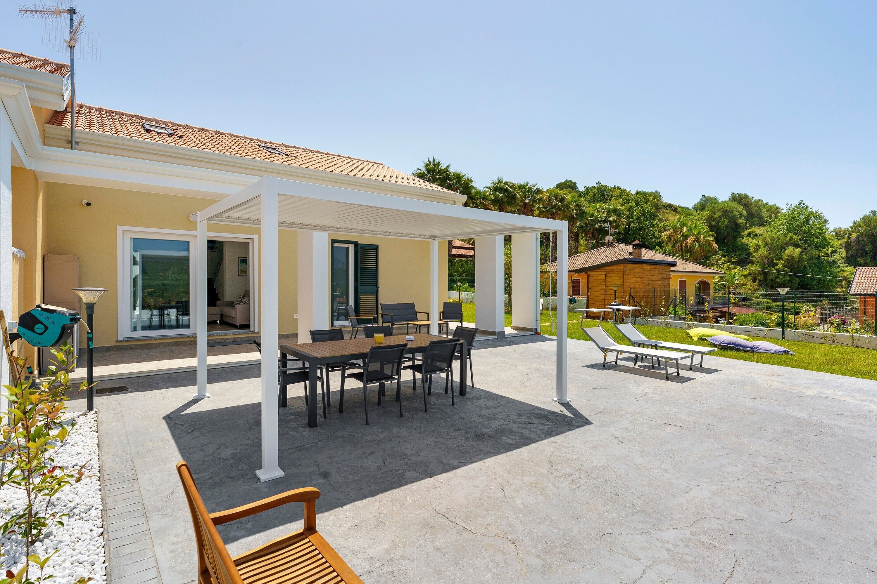 Une terrasse extérieure couverte, avec une table et des chaises, un banc en bois et un espace piscine avec des chaises longues.