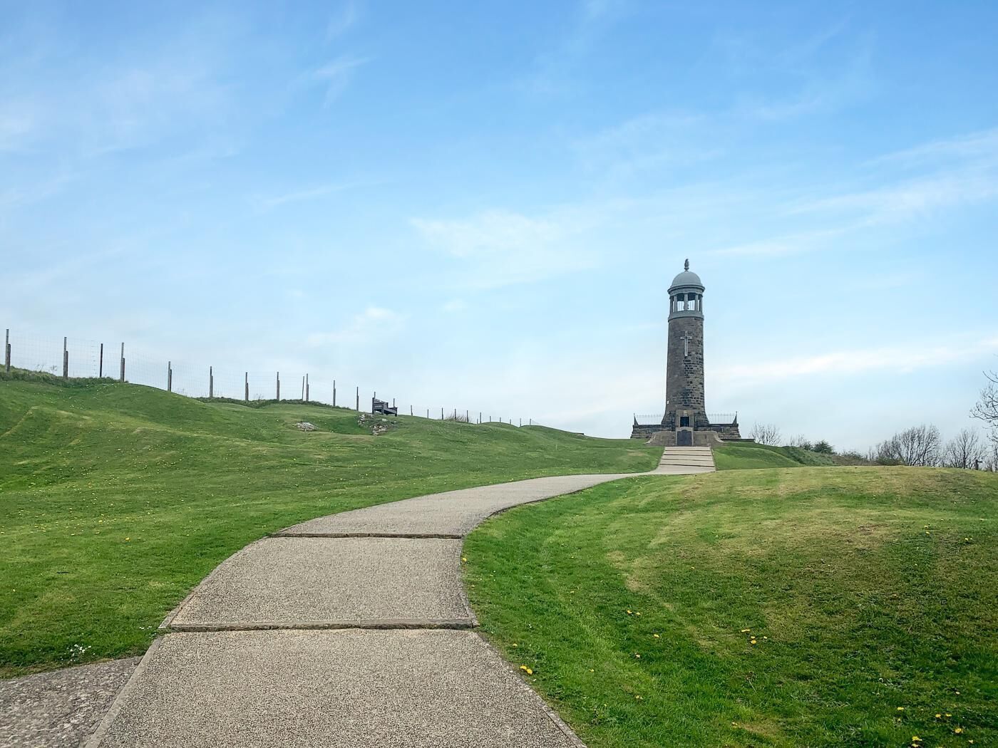 Ein Steinturm mit einem Kreuz, ein gepflasterter Weg dorthin und ein eingezäuntes Gelände mit einem grasbewachsenen Hügel.