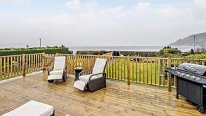 Una terraza de madera con dos sillas y una parrilla, con vista a un área verde y al mar.