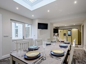 A dining area with a long table set for four, a television mounted on the wall, and a kitchen visible in the background.