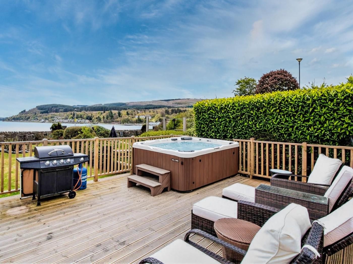 Eine Terrasse mit Whirlpool, Grill und Korbmöbeln mit Blick auf ein Gewässer.