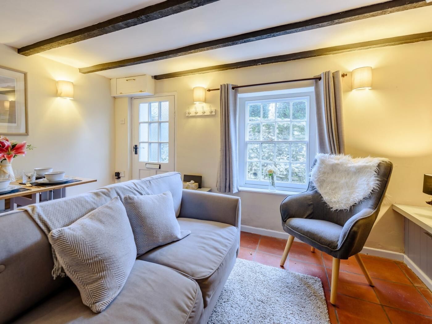 Ein gemütliches Wohnzimmer mit einer Couch, einem Sessel, einem Esstisch mit Geschirr und einem Fenster mit Vorhängen.