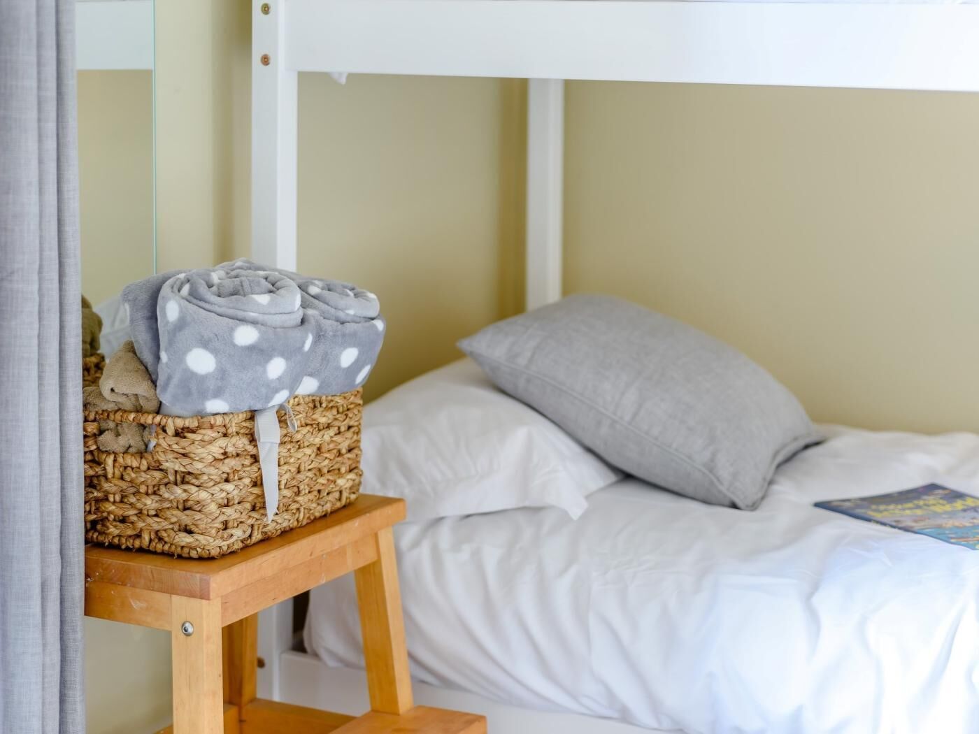 A bed with white bedding, a grey pillow, and a woven basket with folded items on a wooden nightstand.