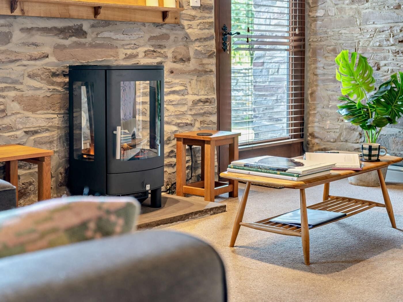 A cozy room with a stone wall, a black wood-burning stove, a wooden table with books, and a potted plant.