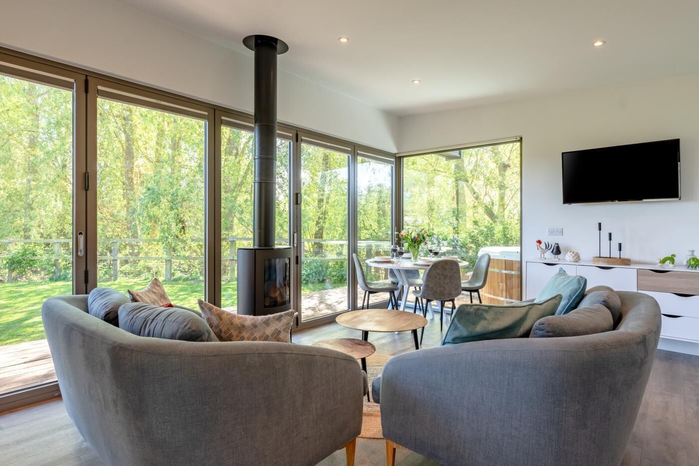 A modern living room with a fireplace, large windows, a dining area, and a flat-screen TV.