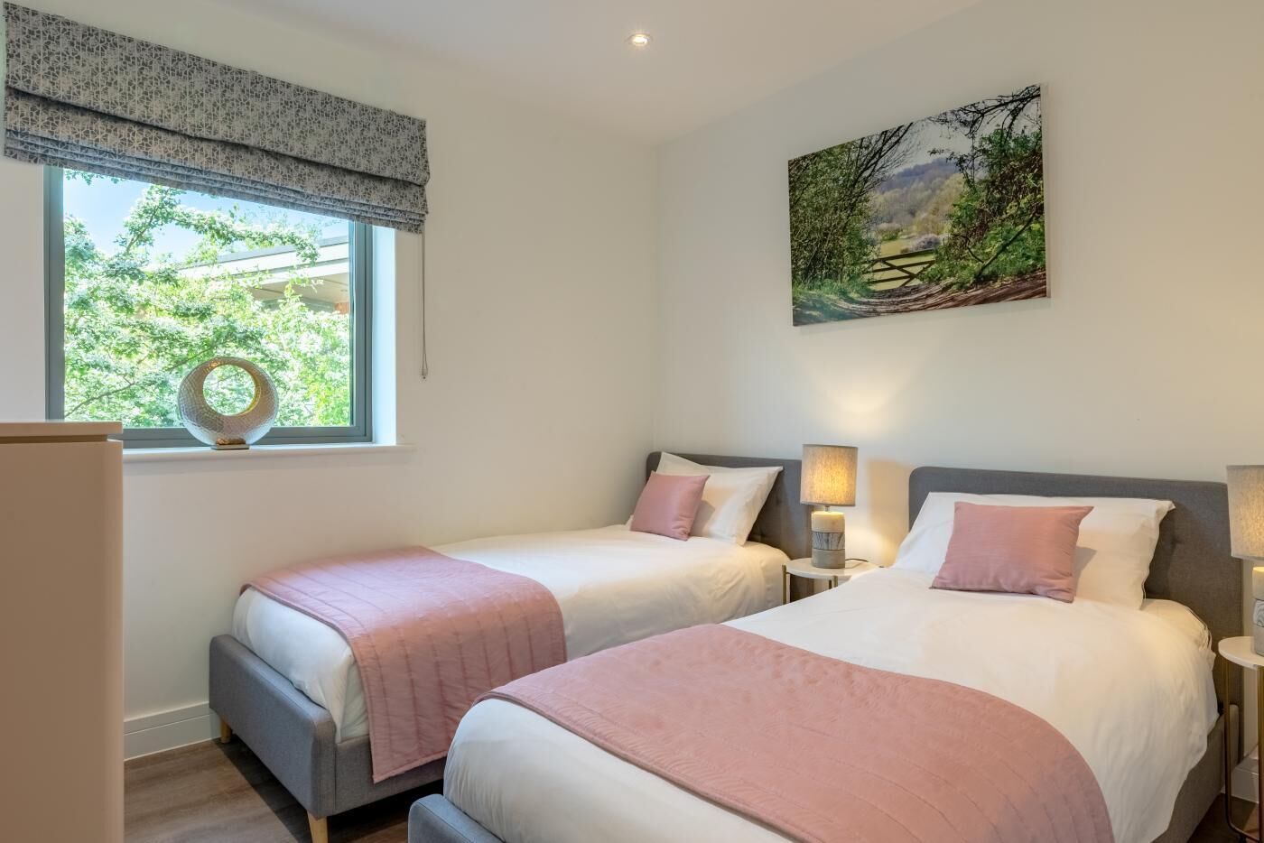 A bedroom with two beds, a window with a view of greenery, a painting on the wall, and a lamp on the nightstand.