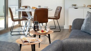 Una sala de estar moderna con un sofá gris, una mesa de centro con una revista, y una mesa para el desayuno con una cafetera y pasteles.