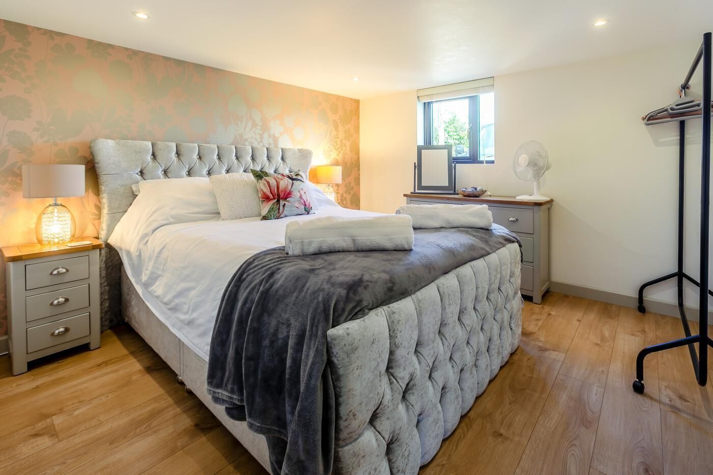 A bedroom with a large bed, bedside tables, a nightstand, a window, and a standing fan.