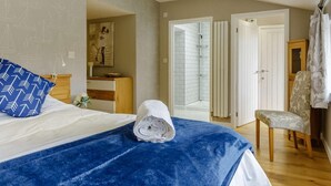 Une chambre avec un lit, une couverture bleue, une serviette enroulée, une chaise, une commode en bois, un tableau, une douche blanche et une porte.