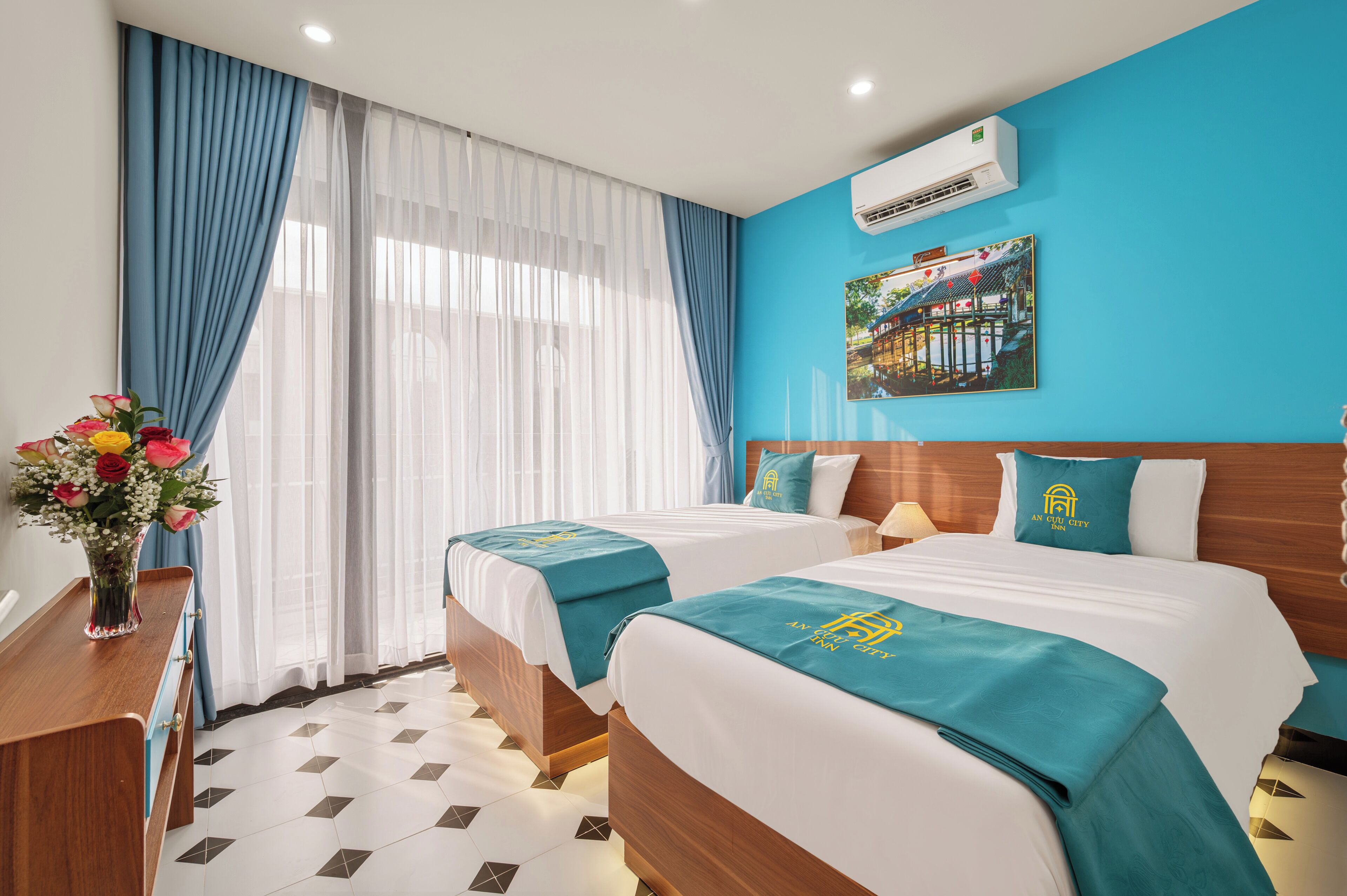 Una habitación de hotel con dos camas, cabecero de madera, aire acondicionado instalado en la pared y una ventana con cortinas translúcidas.