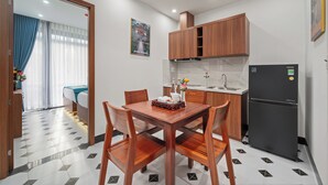 A compact kitchen with wooden cabinets, a refrigerator, and a dining area with a table and chairs. - An Cuu Residence (Hue)