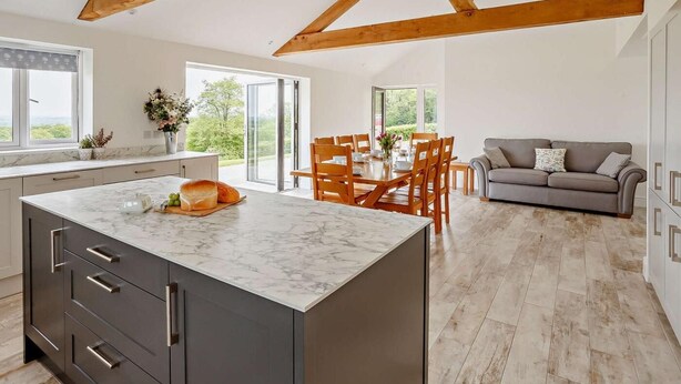Een ruime keuken met een marmeren aanrechtblad, houten balken, een eethoek met een tafel en stoelen, en een grijze bank.