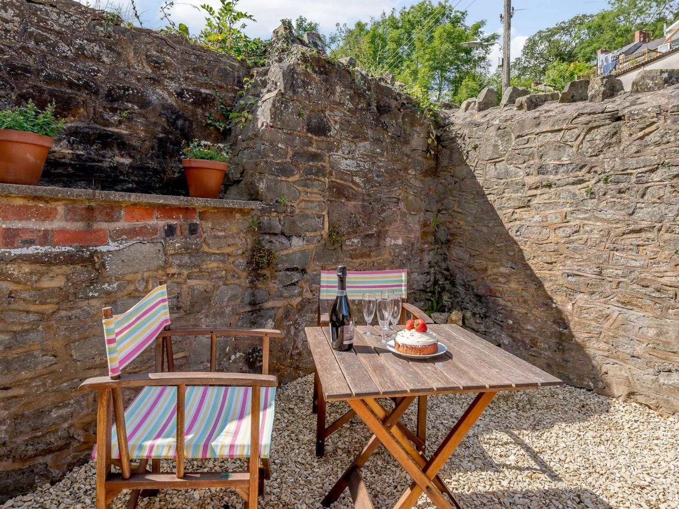 Un espace extérieur rustique aménagé avec une table et des chaises en bois, une bouteille de vin et un gâteau posés sur la table.