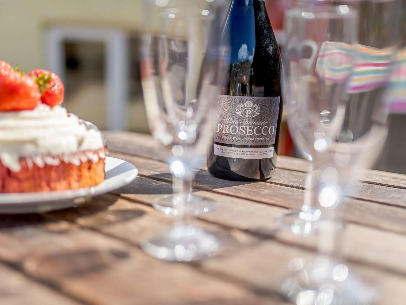 A bottle of Prosecco wine, a plate with a dessert topped with strawberries, and empty wine glasses on a wooden table.