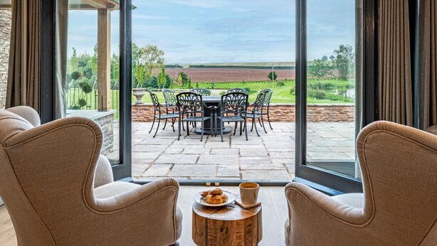 Quarto com uma grande porta de correr de vidro que dá para um pátio exterior com uma mesa e cadeiras, uma pequena mesa redonda de madeira com um prato de comida e uma chávena, e duas poltronas viradas para o pátio.