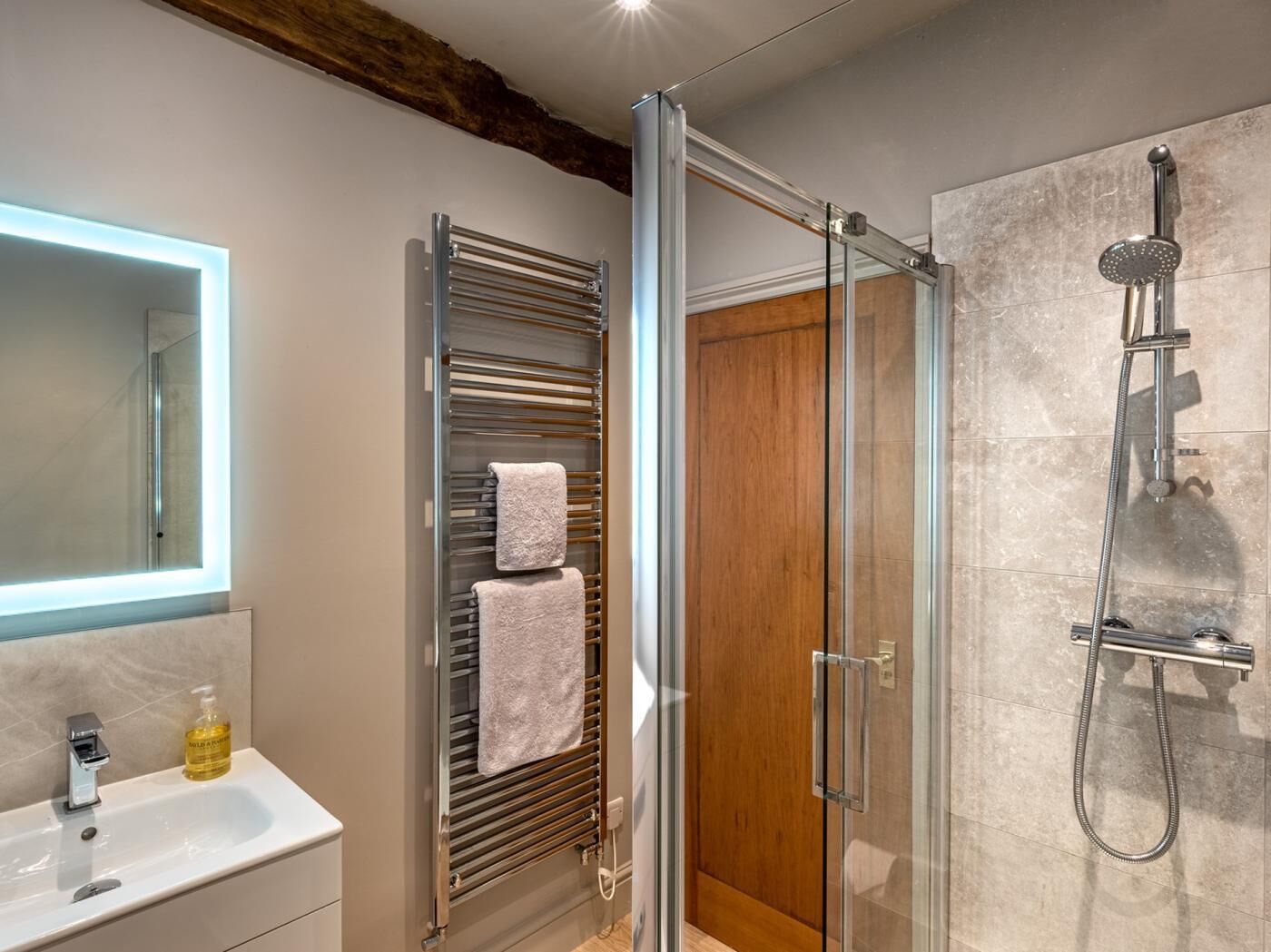 A modern bathroom with a glass shower enclosure, a glass-enclosed towel rack, a wooden door, and a white sink with a soap dispenser.