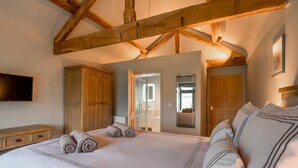 Quarto com uma cama grande, vigas de madeira, um guarda-roupa, uma televisão e um banheiro visível por uma porta aberta.
