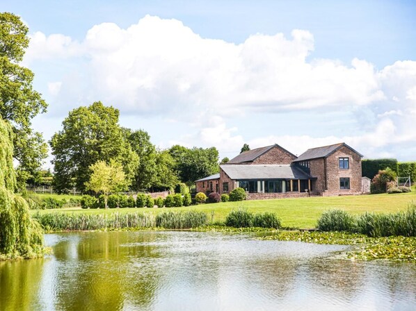 A large brick building with a pond in front, surrounded by lush greenery and trees. - 4 Bed in Hereford (oc-83768) (Saint Weonards)