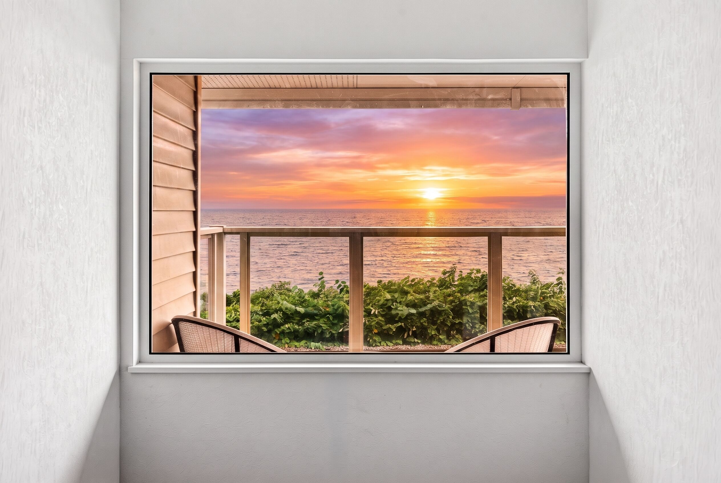 A window with a view of a sunset over the ocean, a balcony with chairs, and a white wall.