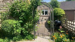 A stone building with a window, a metal archway, and a garden with flowers and a barbecue grill. - 4 Bed in Matlock (oc-85450) (Brassington)