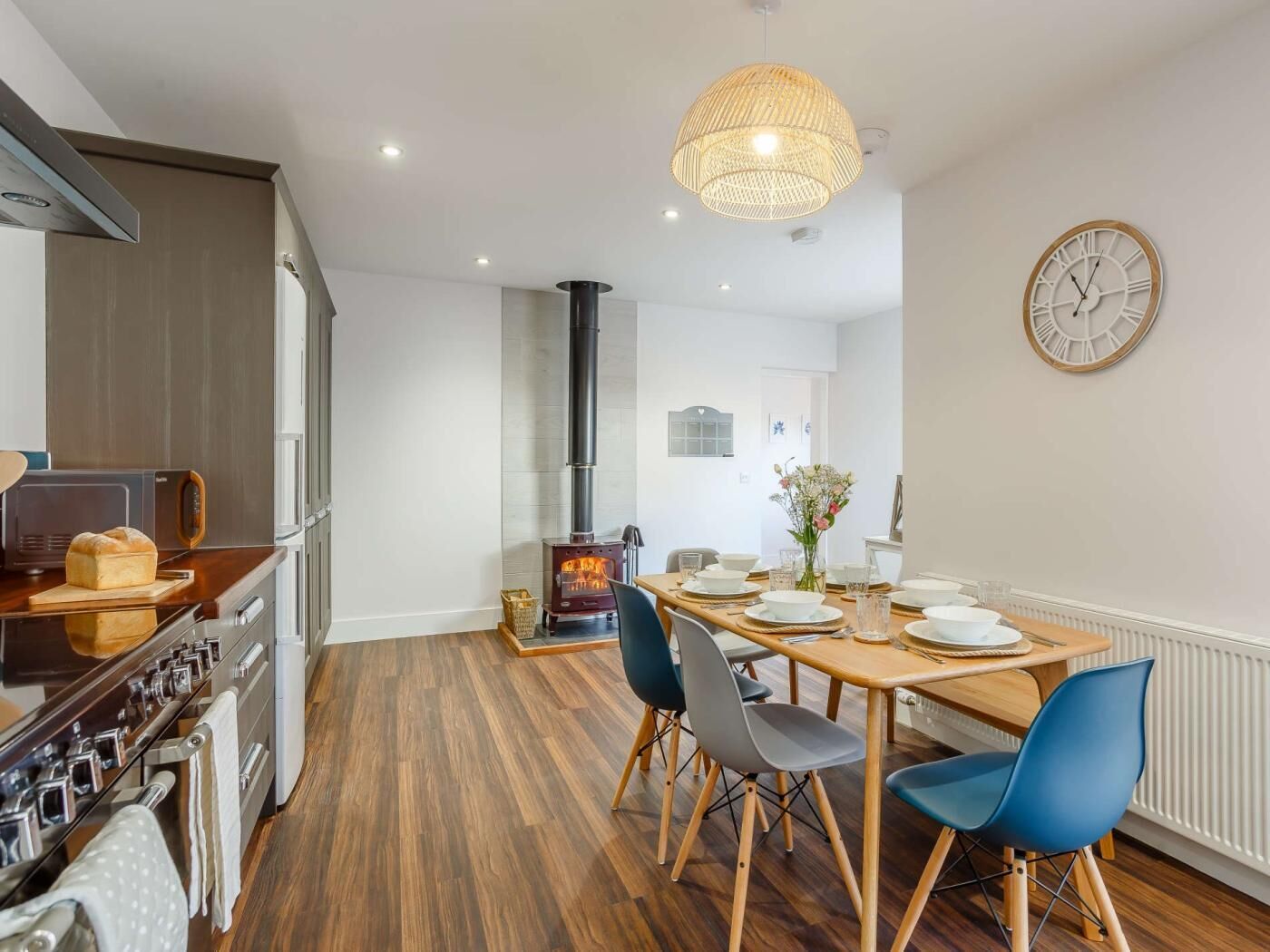 A modern kitchen with a wood-burning stove, a dining area with a wooden table and blue chairs, and a wall clock.