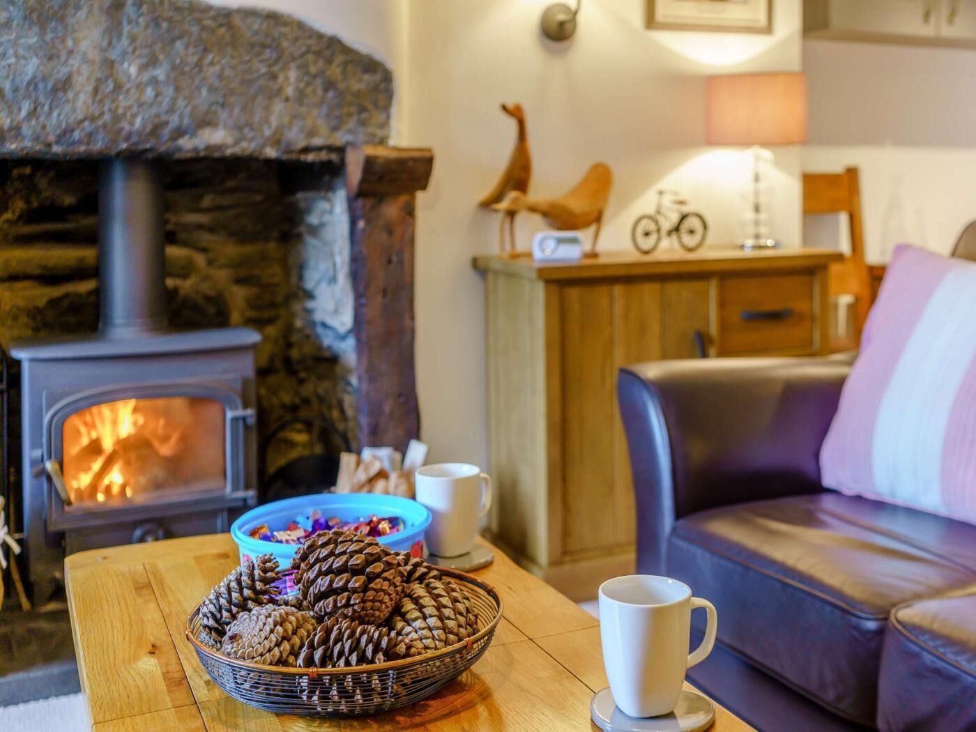 A cozy room with a stone fireplace, a wicker basket of pinecones, a white mug, and a leather sofa.