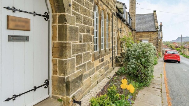 A stone building with a white door, a red car parked on the street, and a garden with flowers. - 3 Bed in Whitby (oc-85927) (Fylingdales)