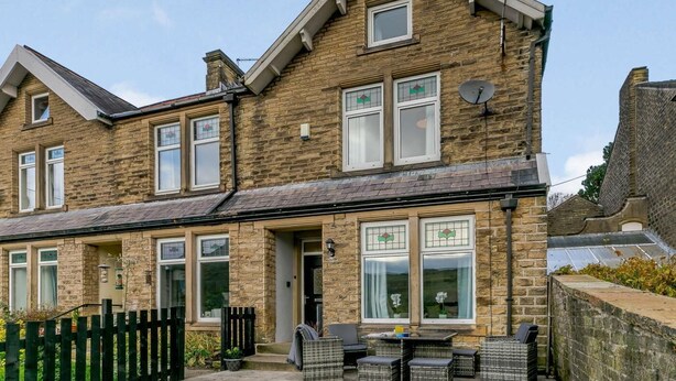 A two-story stone house with a visible front door, a small patio with outdoor furniture, and a satellite dish on the roof. - 4 Bed in Holmfirth (oc-86032) (Holmfirth)
