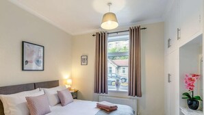 A bedroom with a bed, a bedside lamp, a window with curtains, a radiator, and a picture on the wall. - 4 Bed in Holmfirth (oc-86032) (Holmfirth)