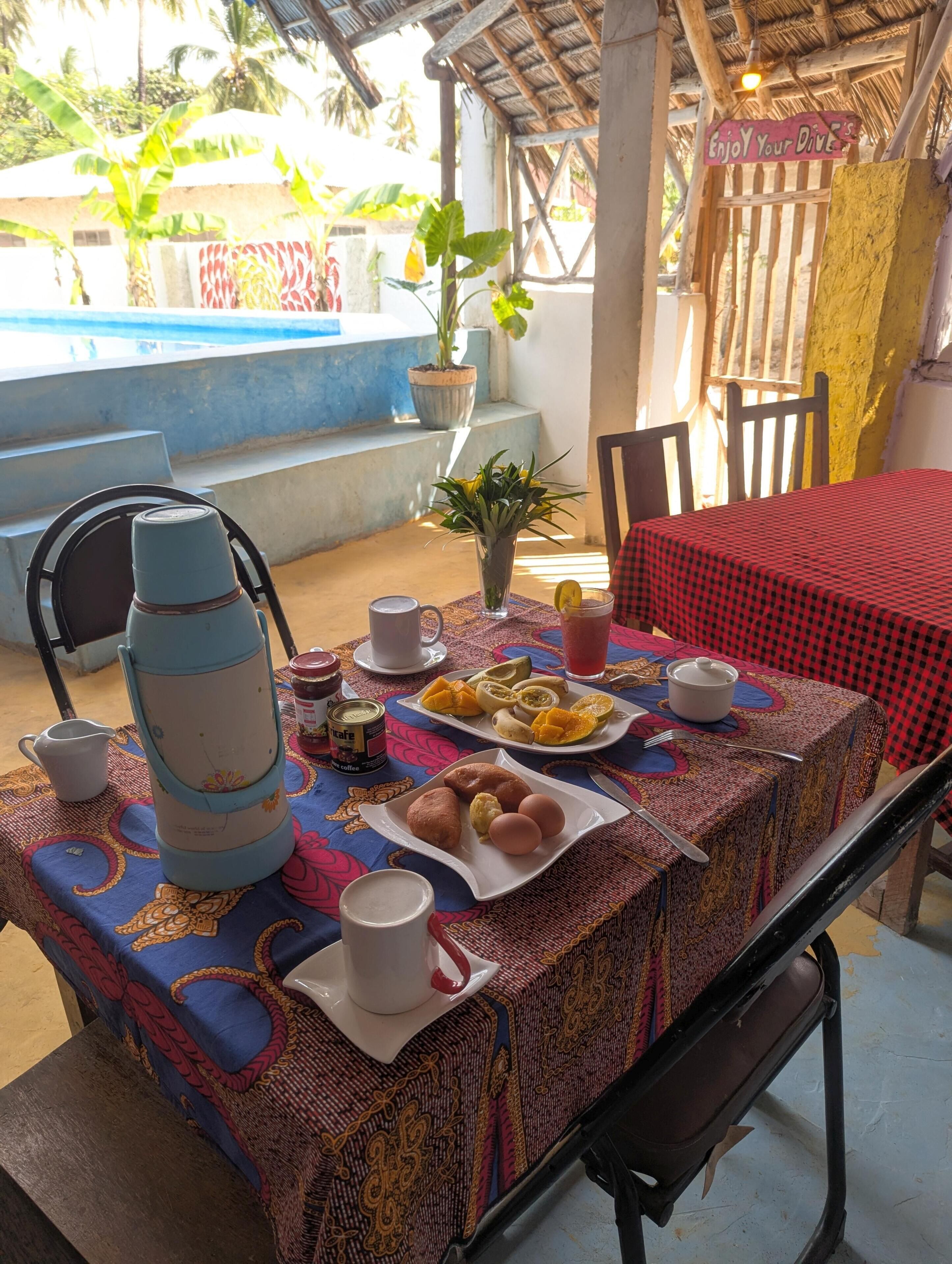Mesa de jantar posta com diversos alimentos e bebidas, uma cafeteira, e uma placa com a inscrição 'Aproveite os seus mergulhos'.