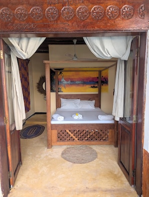 A wooden bed with a headboard, white curtains, and a painting of a sunset.