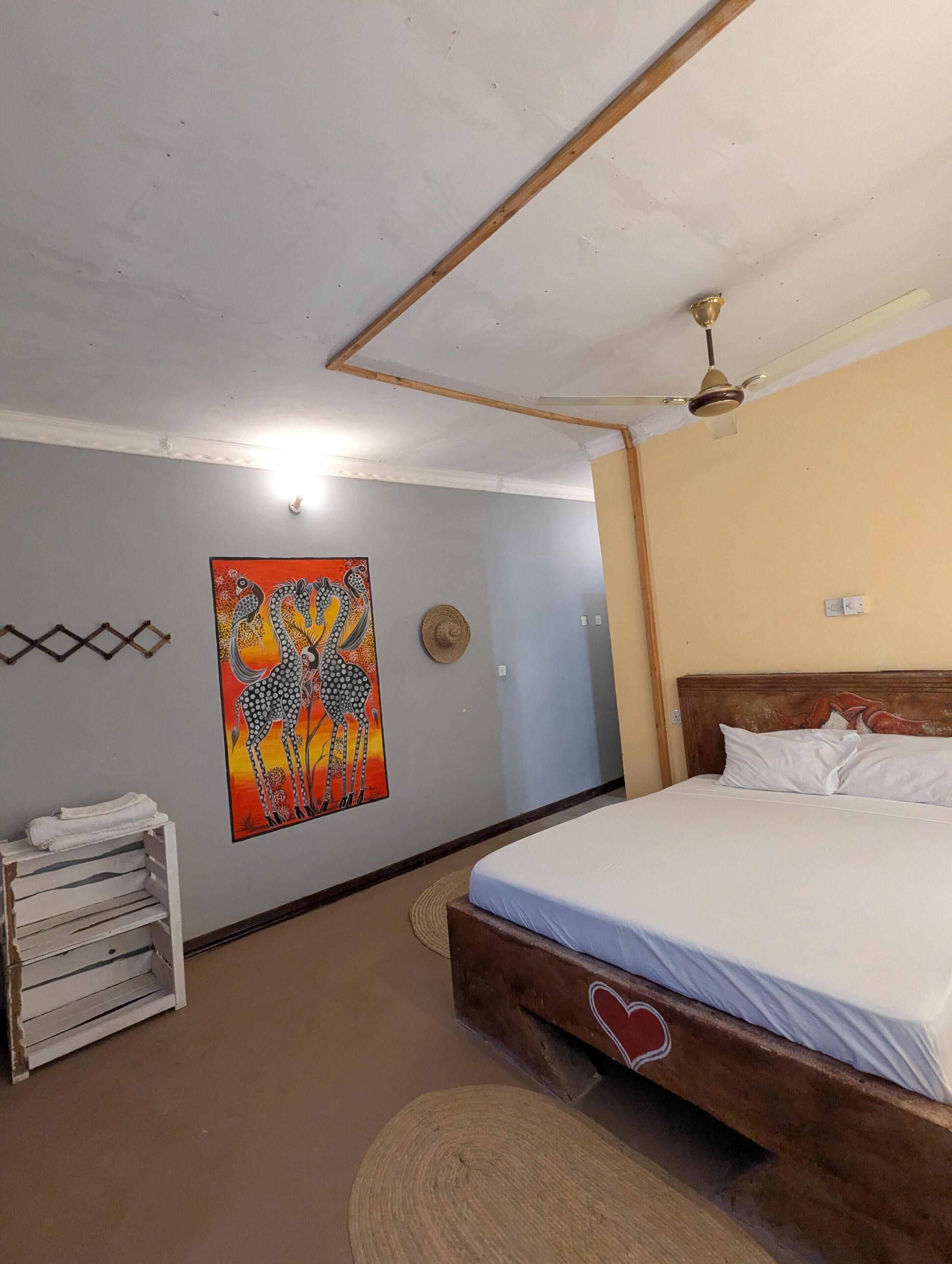 A bedroom with a bed, a wall-mounted heater, a ceiling fan, and a painting of giraffes.