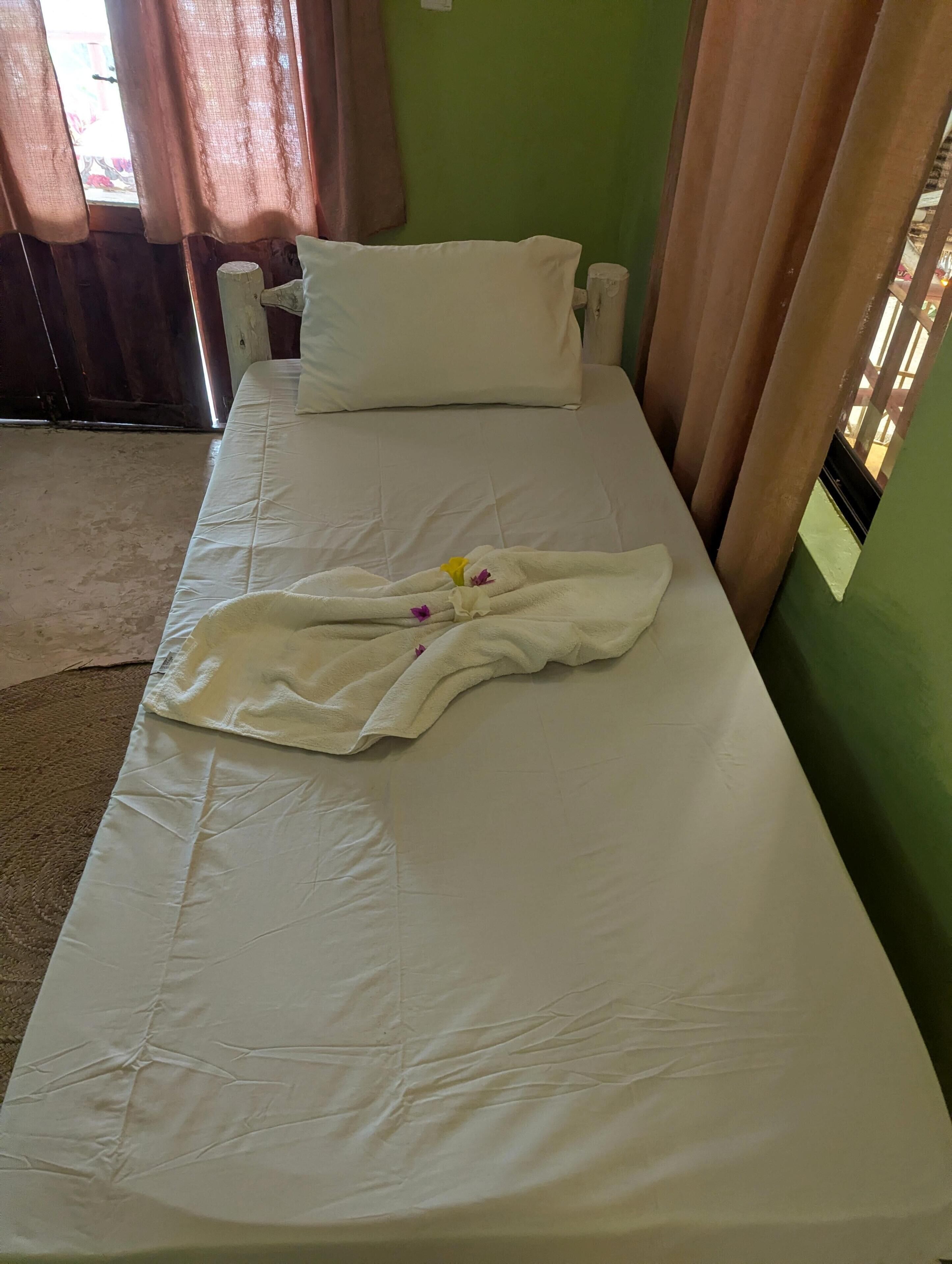 Habitación con cama individual, colcha blanca, almohada y toalla con flores.