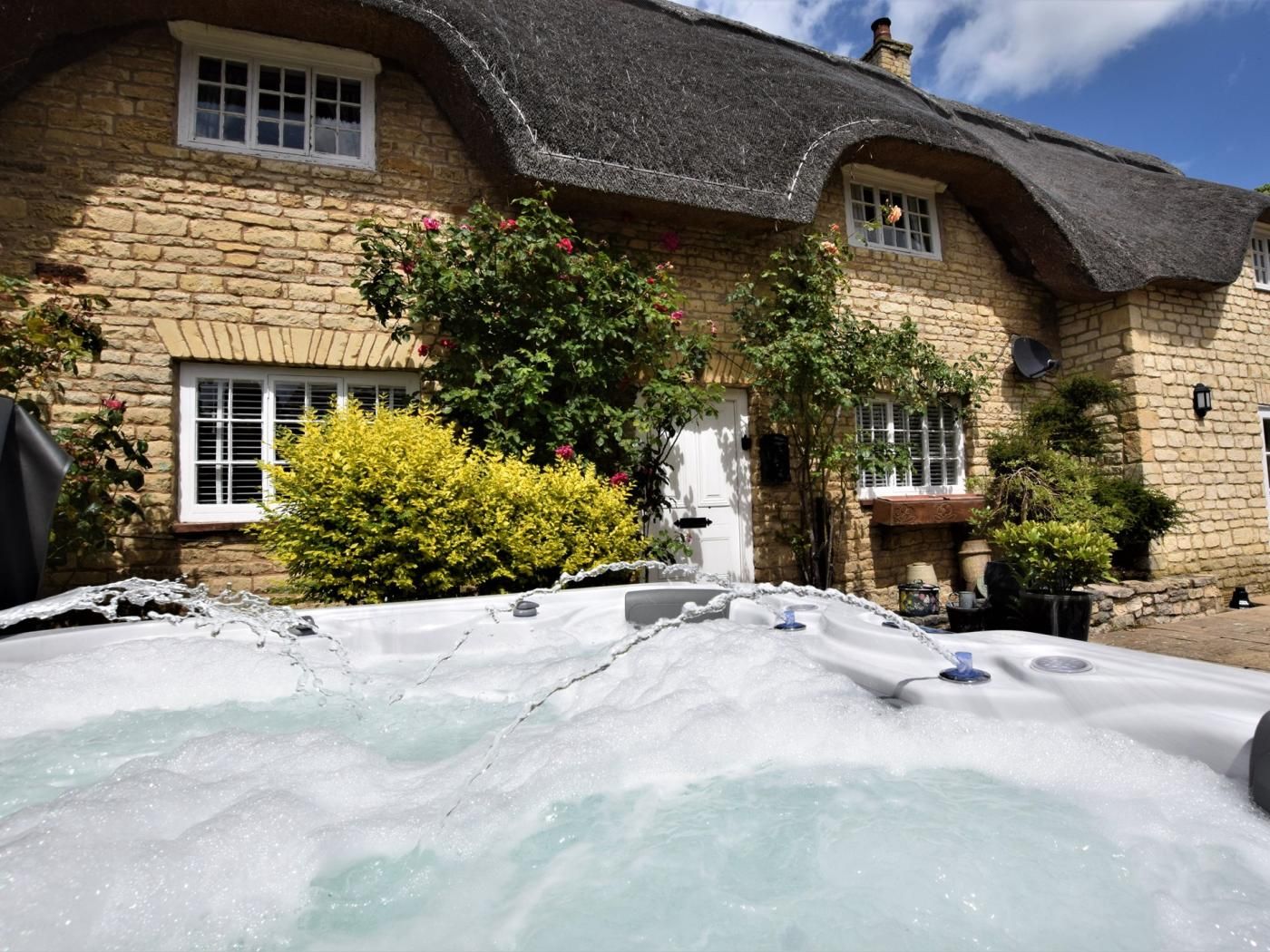 A stone house with a thatched roof, a hot tub, and lush greenery.