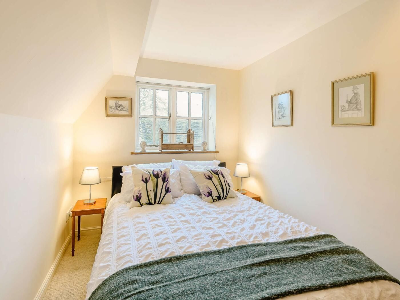 A bedroom with a bed, two bedside tables, a window, and three framed pictures on the wall.