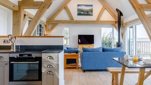 Uma cozinha moderna com forno, área de jantar com mesa e sofá azul, e televisão em um quarto estilo loft com vigas de madeira e uma pintura.