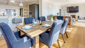 Una cocina moderna con armarios blancos, un comedor con mesa de madera y sillas azules, y una sala de estar con televisor y chimenea.