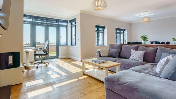 Una sala de estar moderna con un sofá, una mesa de centro y un escritorio con una silla. Amplias ventanas dejan entrar luz natural y ofrecen vistas al exterior.