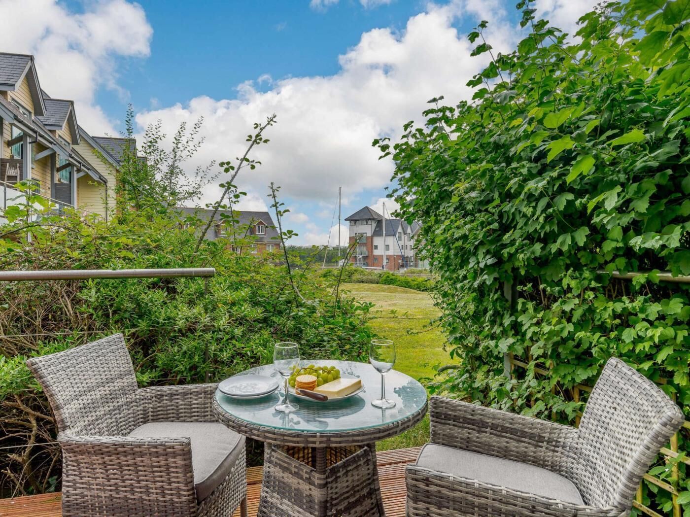 A wicker outdoor seating area with a table set for two, a grassy lawn, and houses in the background.