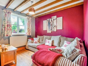 A cozy room with a wooden ceiling, a sofa with floral cushions, a red throw blanket, and a window with floral curtains.