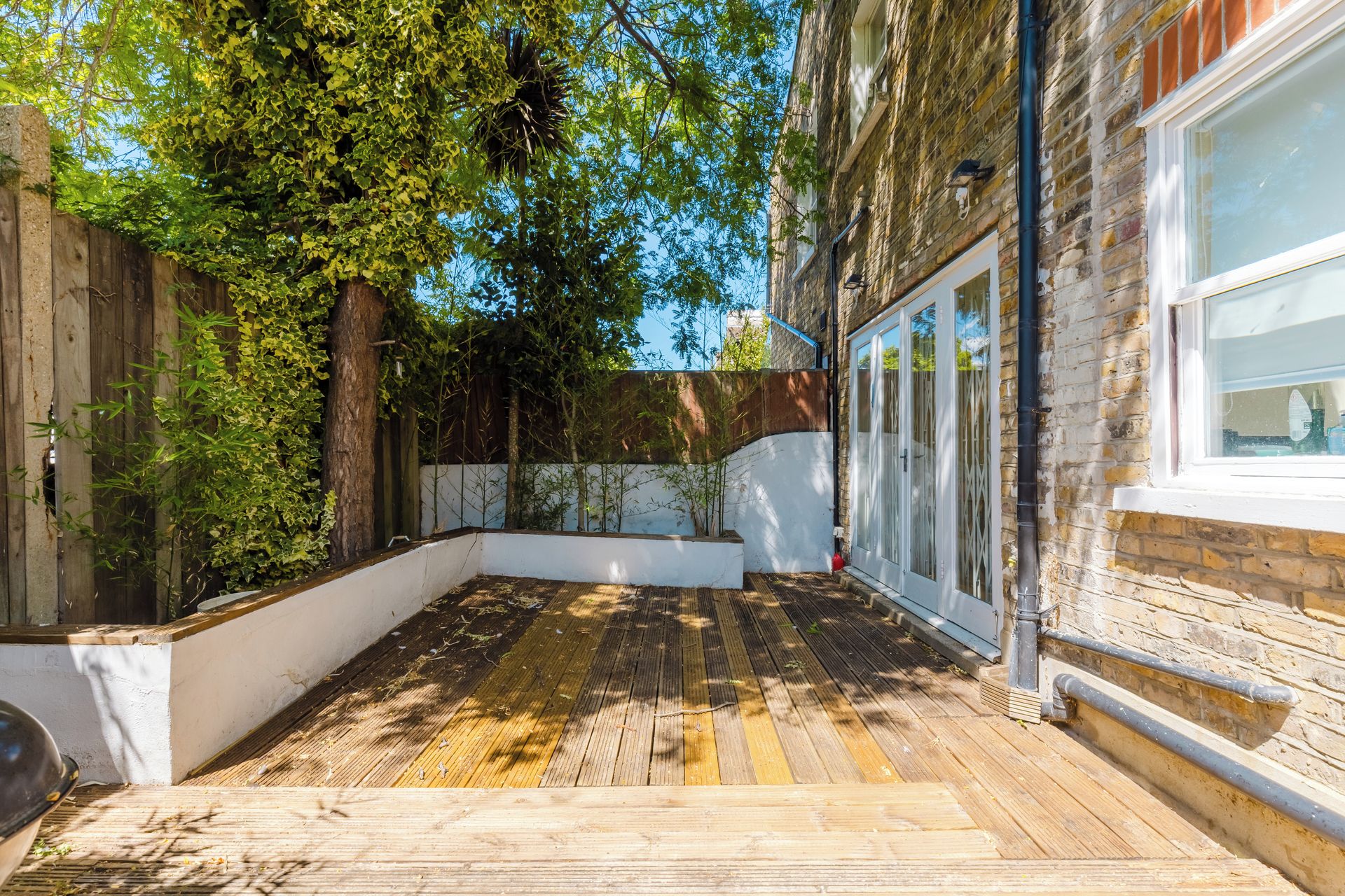 Une terrasse en bois avec un bac à fleurs blanc, un mur de briques et une fenêtre avec des barreaux en métal.