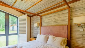 Un dormitorio con techo de madera, una cama con ropa de cama blanca, una mesita de noche, una lámpara y vistas al exterior.