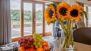 Ein Esstisch mit Obstschale, einer Vase mit Sonnenblumen und Blick auf eine Brücke und Grünflächen durch große Fenster.