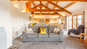 Ein Wohnzimmer mit grauem Sofa, Holzbalken, Heizkörper und Blick auf die Küche sowie den Außenbereich.