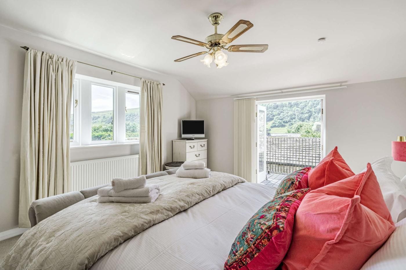 A bedroom with a bed, a television, a ceiling fan, and a view of the outdoors.