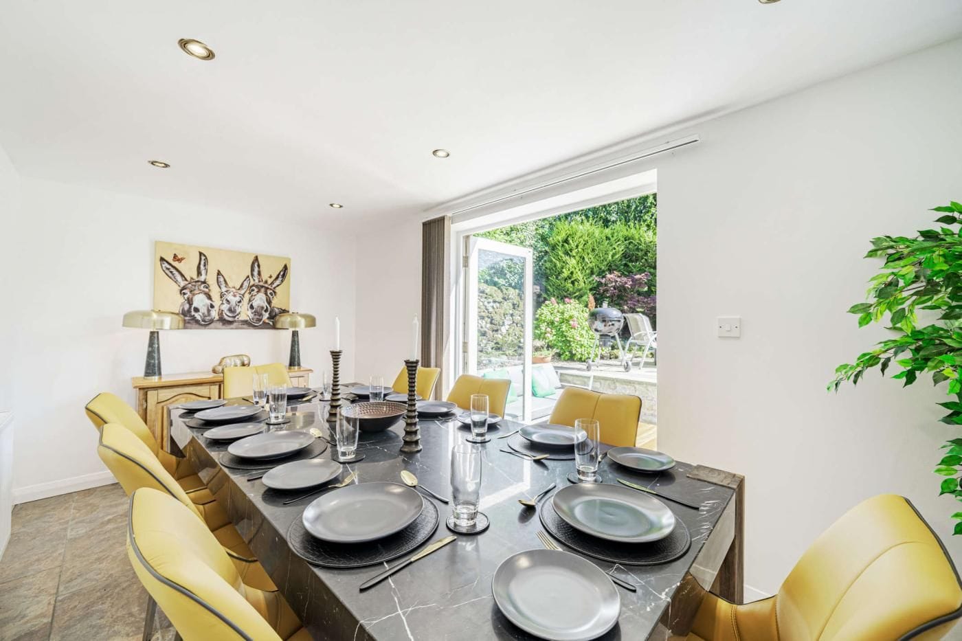A dining area with a long table set for six, yellow chairs, and a painting on the wall.