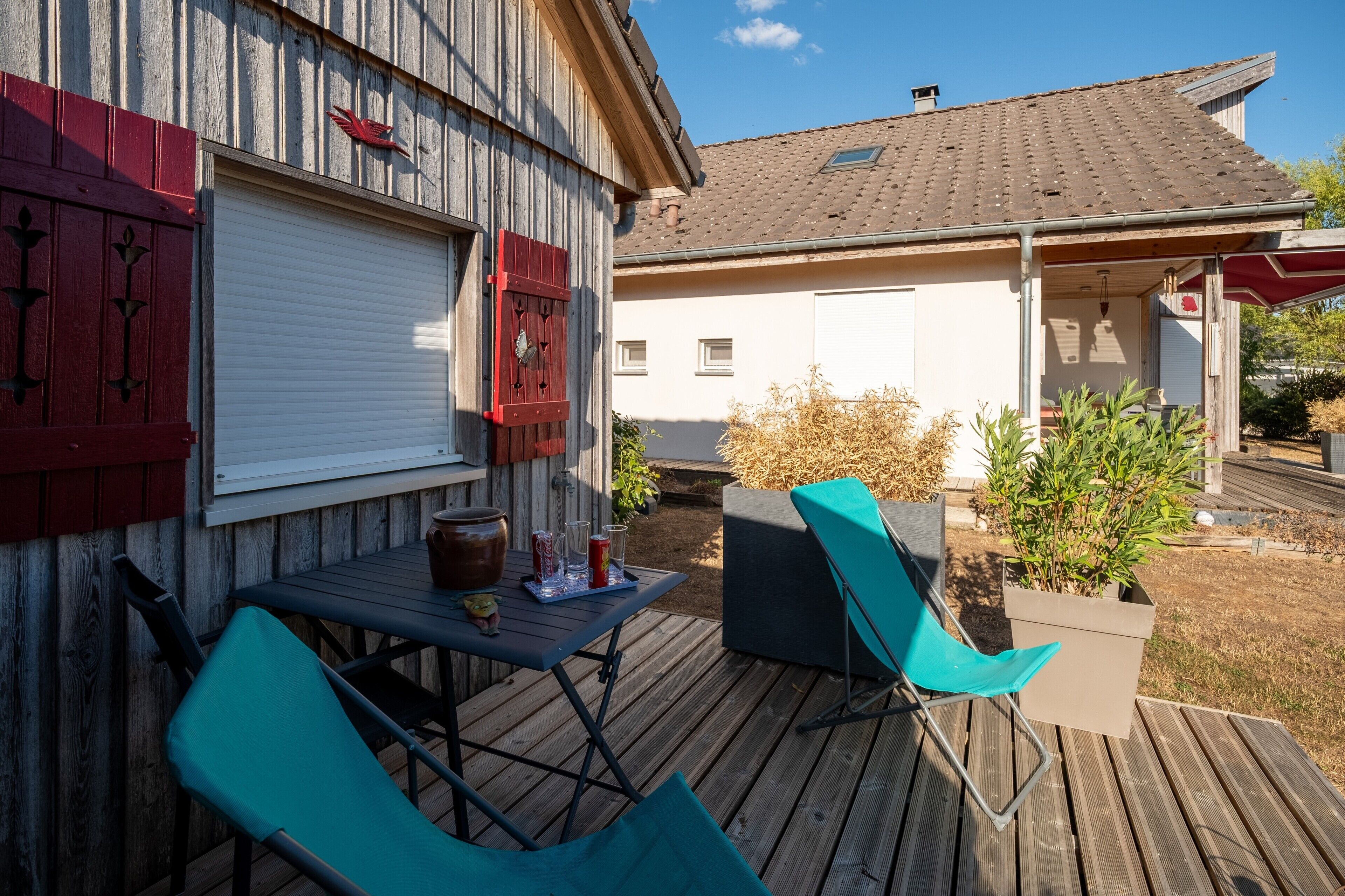 Une terrasse en bois avec une table et des chaises, une boîte aux lettres rouge et un bâtiment blanc en arrière-plan.